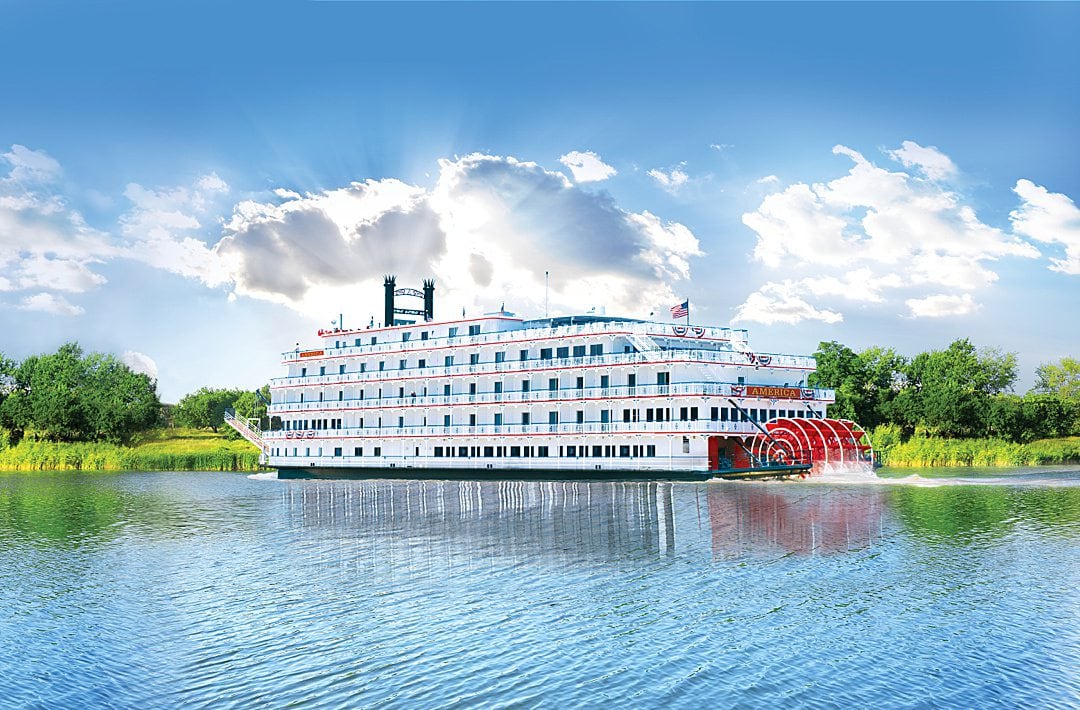 American Cruise Lines US river cruise in the water with greenery behind it and blue skies and sunshine streaming through the clouds.