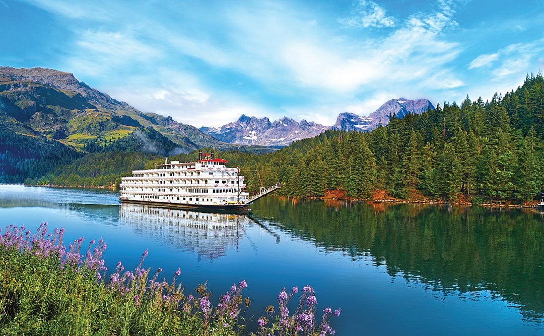 American Cruise Lines US river cruise in the water with mountains behind it and purple flowers in the foreground.