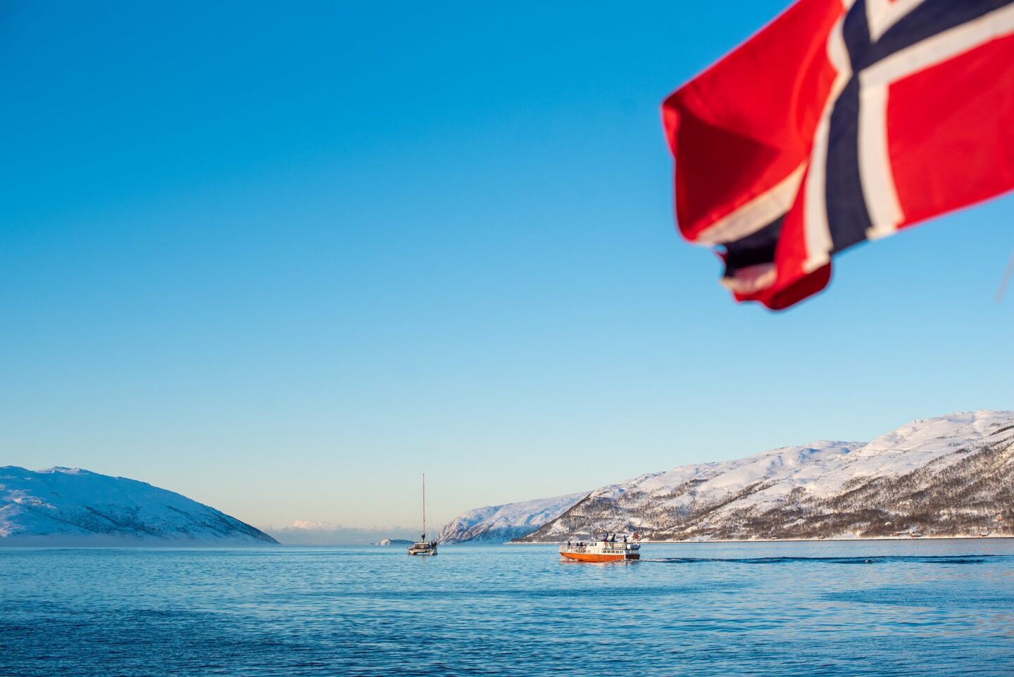 Norwegian flag flying in the top right corner on a Polar Fjord Cruise with the snowy mountains and blue sky and arctic ocean in the background.