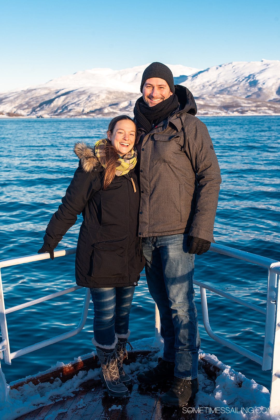 A couple at the stern of a ship on a Tromso, Norway boat tour with snowy mountains in the background.