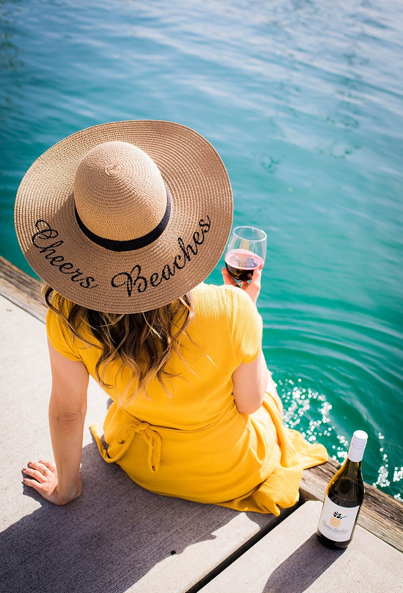 Woman's back in a yellow dress with a custom hat that says Cheers Beaches, holding a glass of red wine.