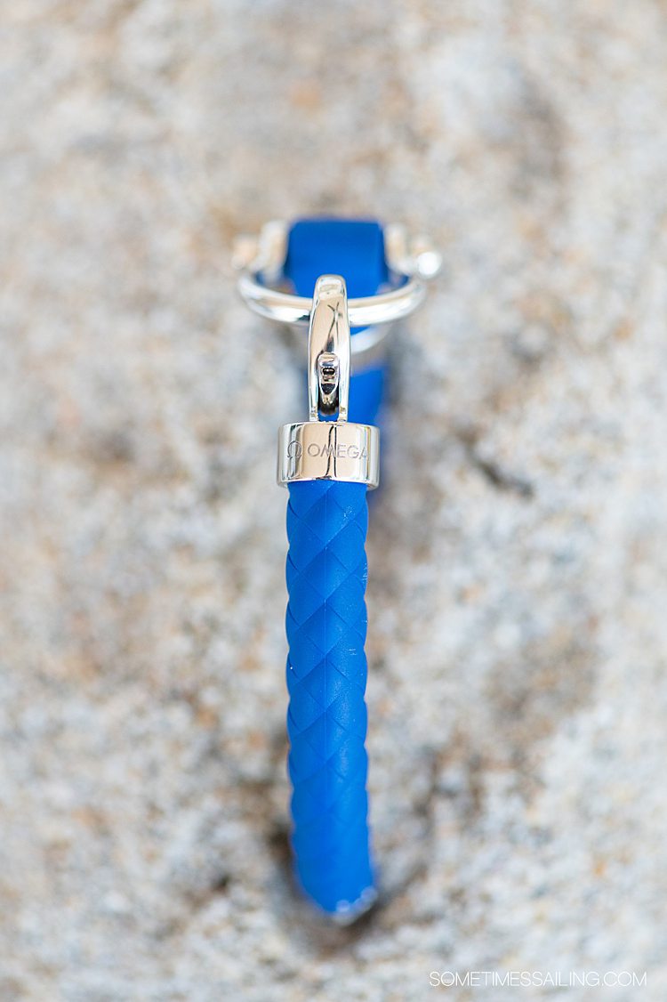 Blue rubber and stainless steel sailing bracelet from OMEGA against sand on the beach and a piece of white coral.