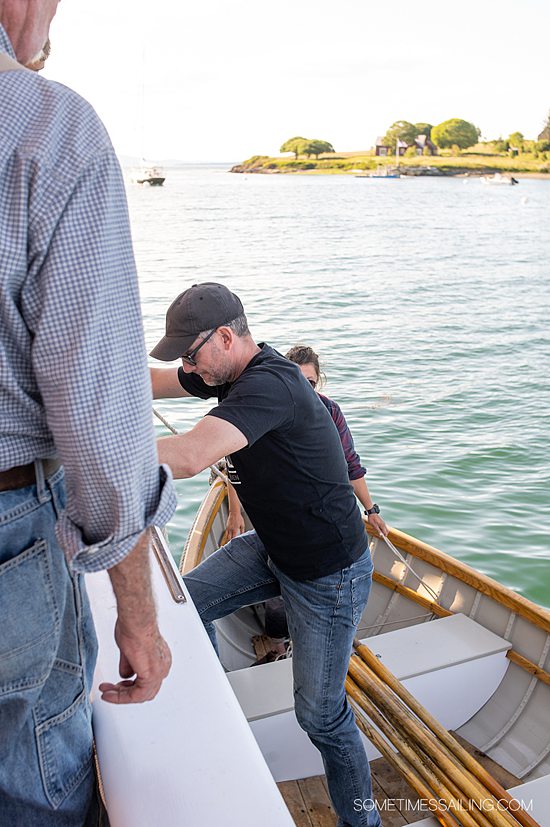 Man going into a rowboat from a sailboat on the water in Maine.