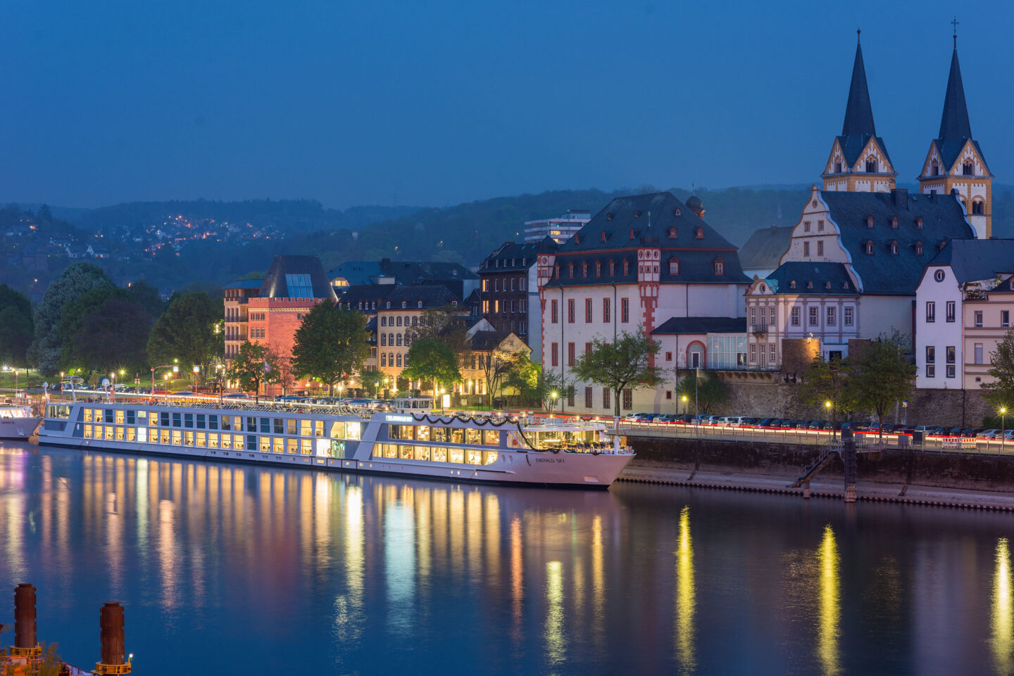 River cruise ship in port in Europe, with the "Emerald Sky" words on the side of the ship and the night sky, and lights reflecting in the river.