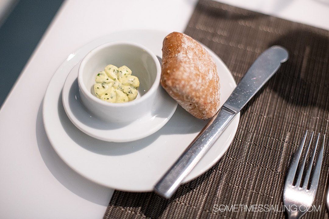 Plate with a roll and butter knife on the right, and small white ramekin filled with herb butter.