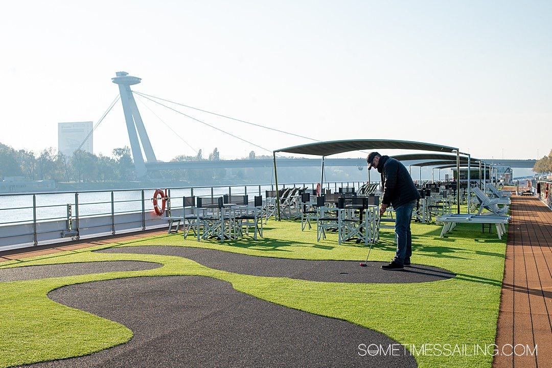 Man dressed in black playing putt-putt golf on the top deck of a river cruise ship, with a bridge in the background.