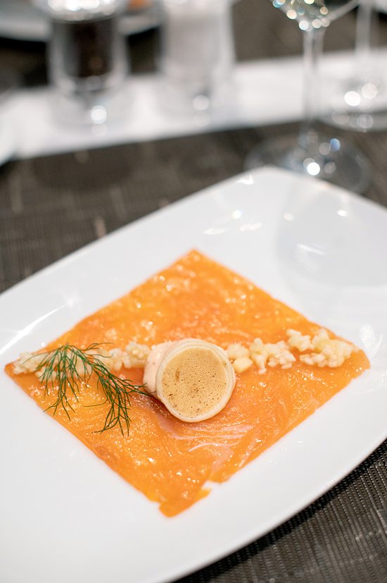 Plate of smoked salmon arranged in a cut square shape, with a tiny pancake on top and dill with a crunchy crumble. 