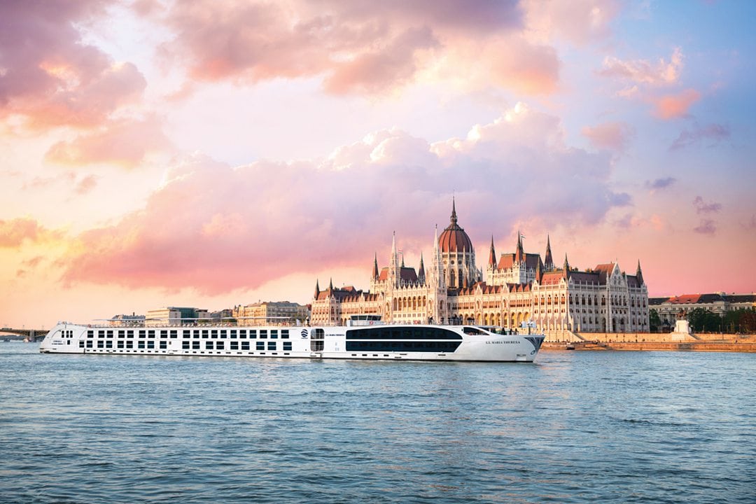 Uniworld river cruise boat in front of the parliament building on the Danube River in Budapest, with a pink and purple sunset sky. 