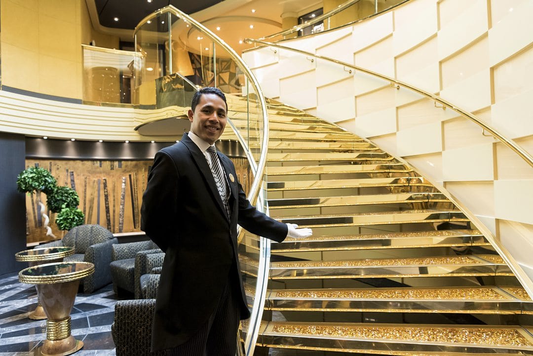 Man in a tuxedo motioning to a luxurious staircase, on MSC Cruises Yacht Club, a ship-wtihin-a-ship cruise experience.