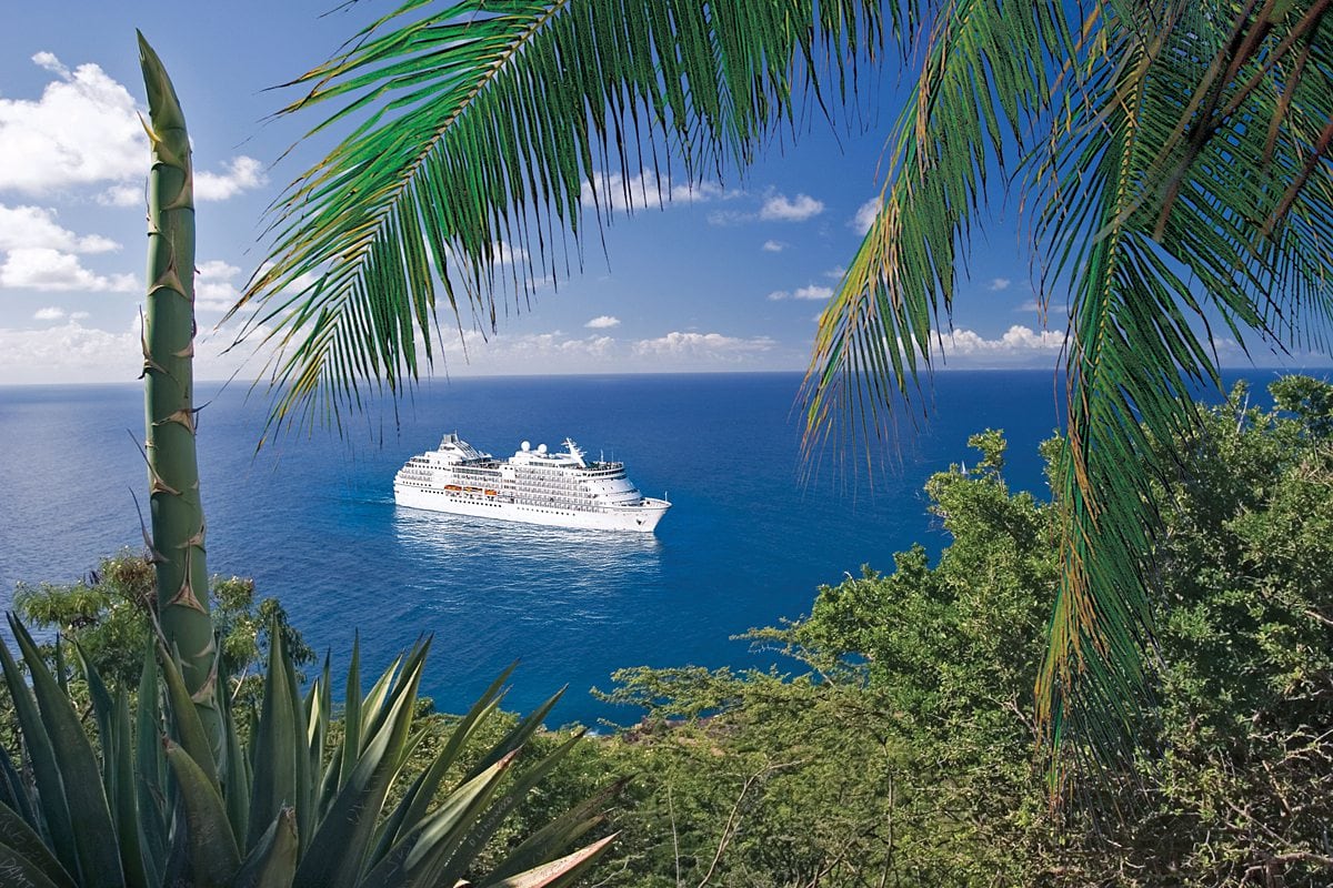 Regent Seven Seas cruise ship in the tropics with blue skies, a blue ocean and palm tree and greenery in the foreground.