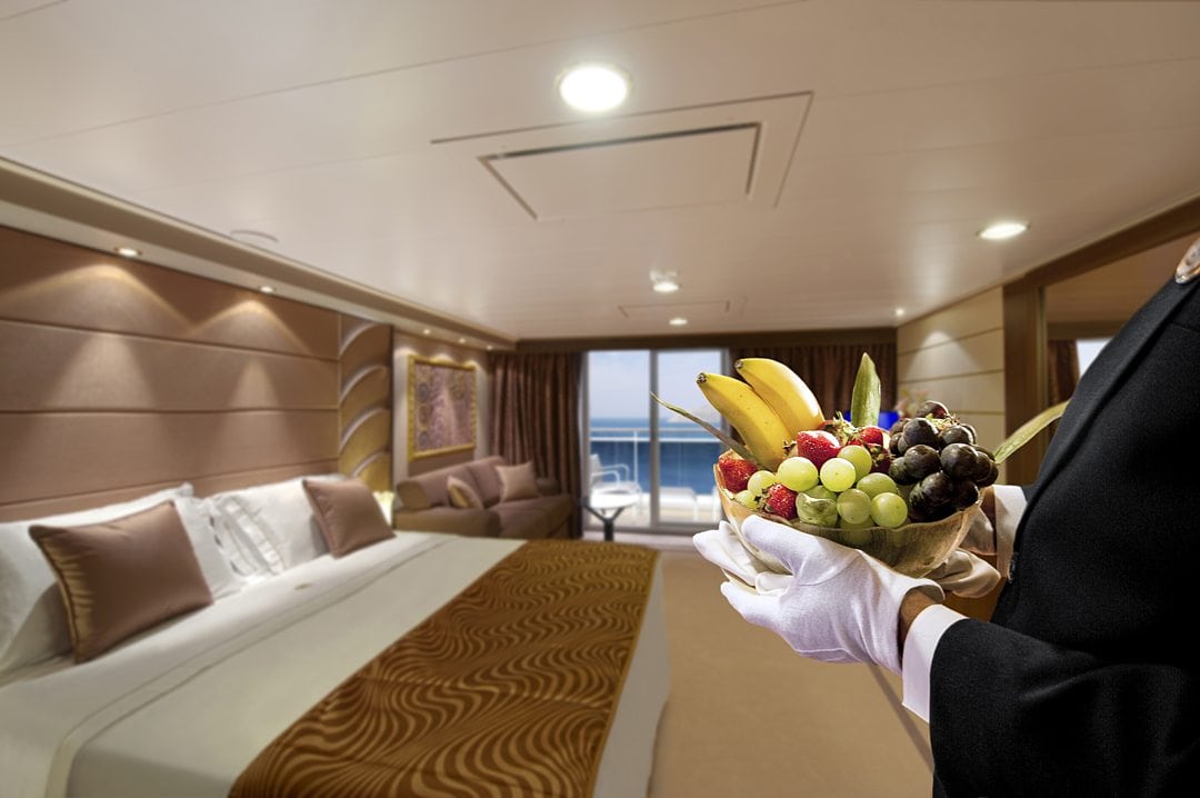 White-gloved hand on the right serving a bowl of fruit with a cruise cabin in the background.