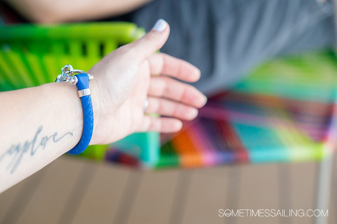 Blue Omega sailing bracelet on a wrist with a colorful background. Wrist has an "explore" tattoo on it.