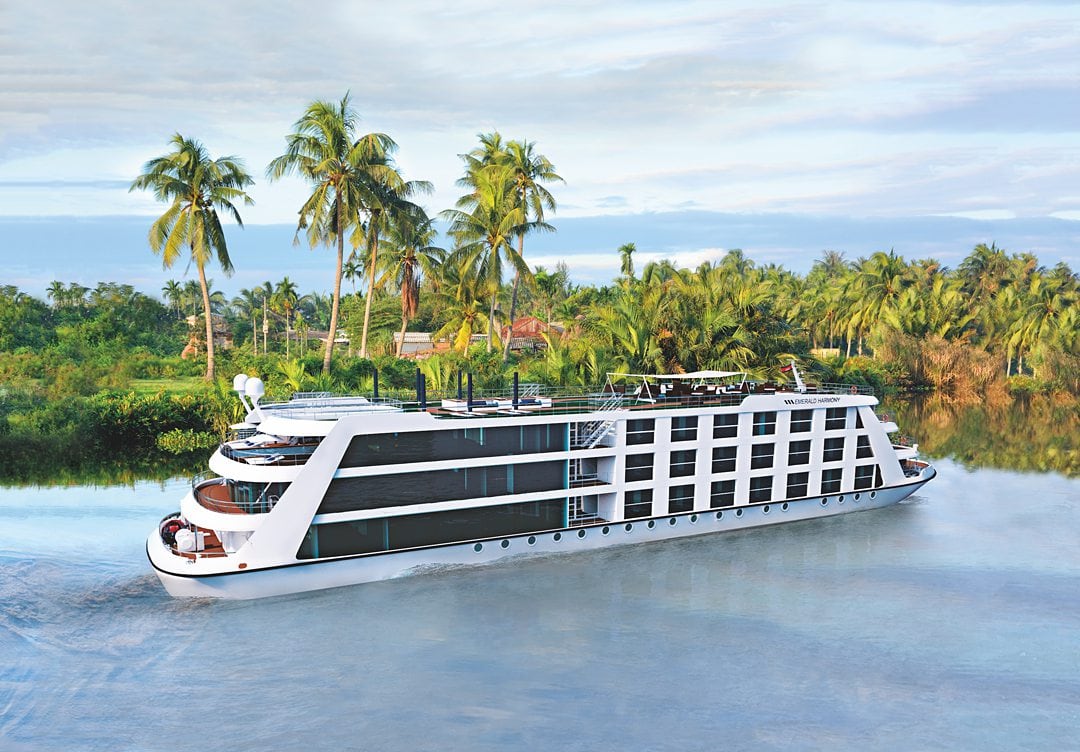 Emerald Harmony on the Mekong River with palm trees behind it and a cloudy blue sky.