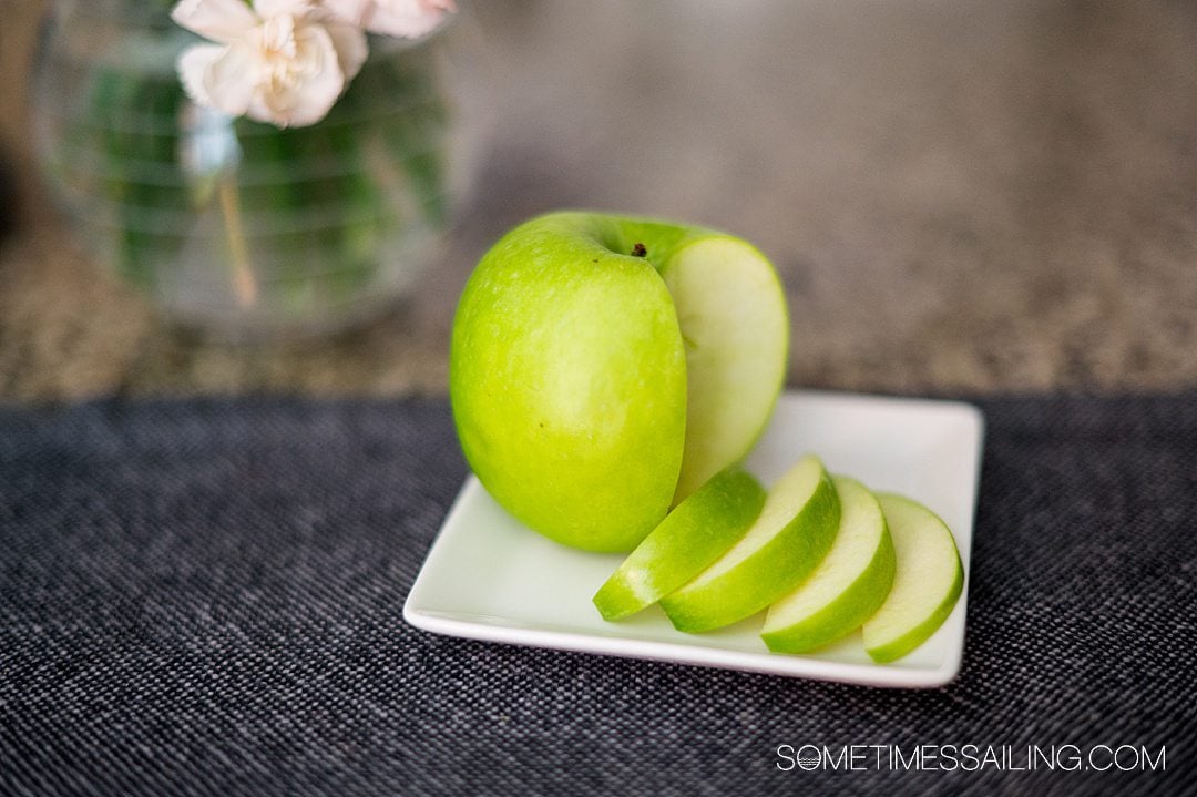 Large green apple on a small square white plate with a quarter cut out and four slices next to it for an anti-nausea remedy on cruises.