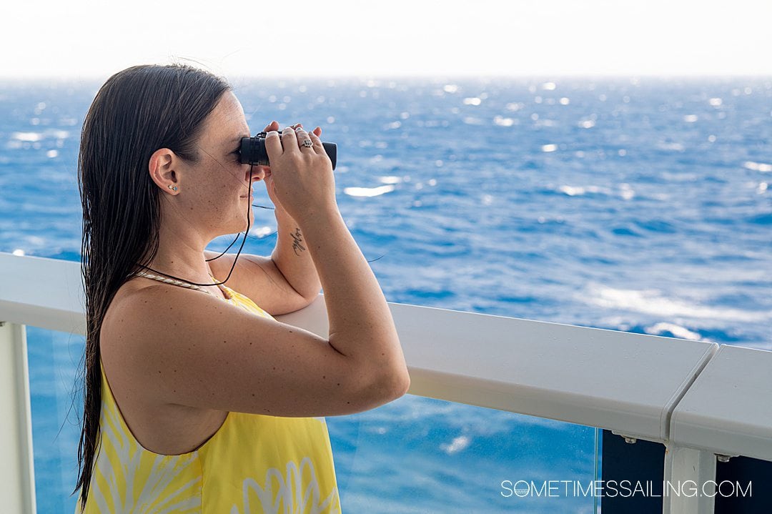 Woman looking through binoculars with the ocean behind her, wearing a yellow shirt.