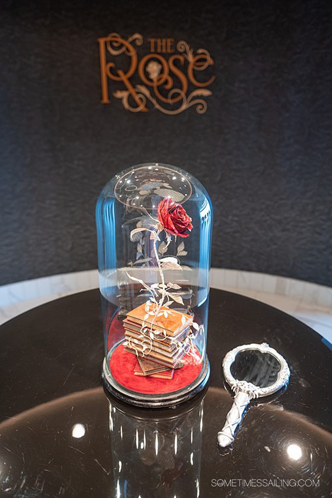 Entrance to The Rose bar on Disney Wish, with a glass encased rose on a black table, with a mirror next to it.