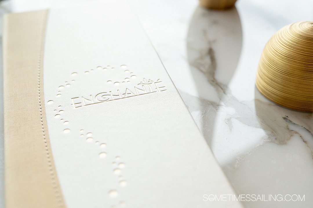 White menu with the word "Enchante" on the front with gold bubbles, resting on a white marble table. This is the wine menu for the restaurant of the same name on Disney Wish cruise ship.