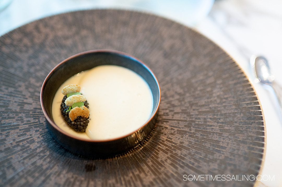 Small, round, flat dish with a cream mixture in it and a dollop of caviar on the left, with three small round crackers in it.