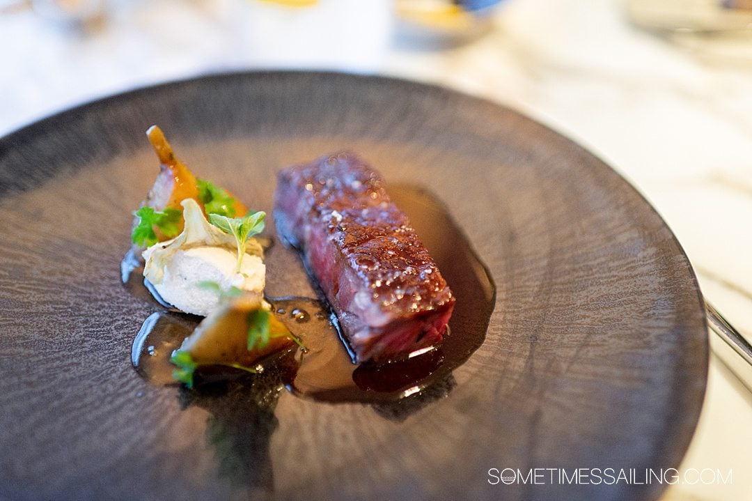 Rectangular cut of meat on a black plate with artichokes on the left and green herbs.