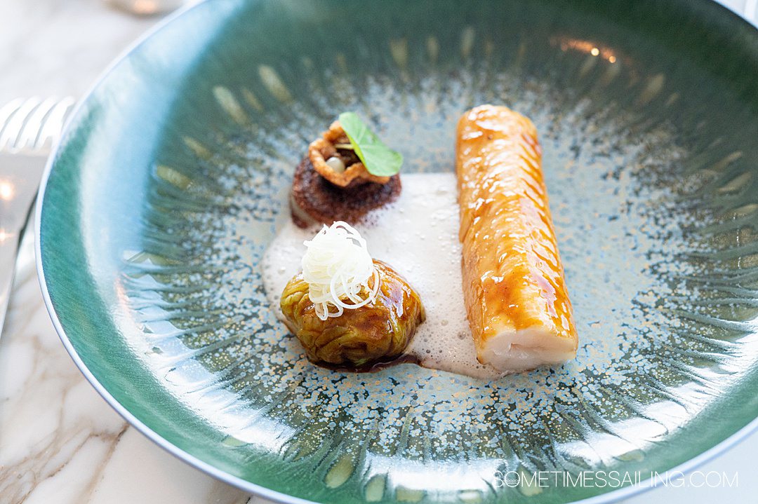 Teal plate with three piece of food on it in a white foam sauce on Disney Wish, at Enchante restaurant.