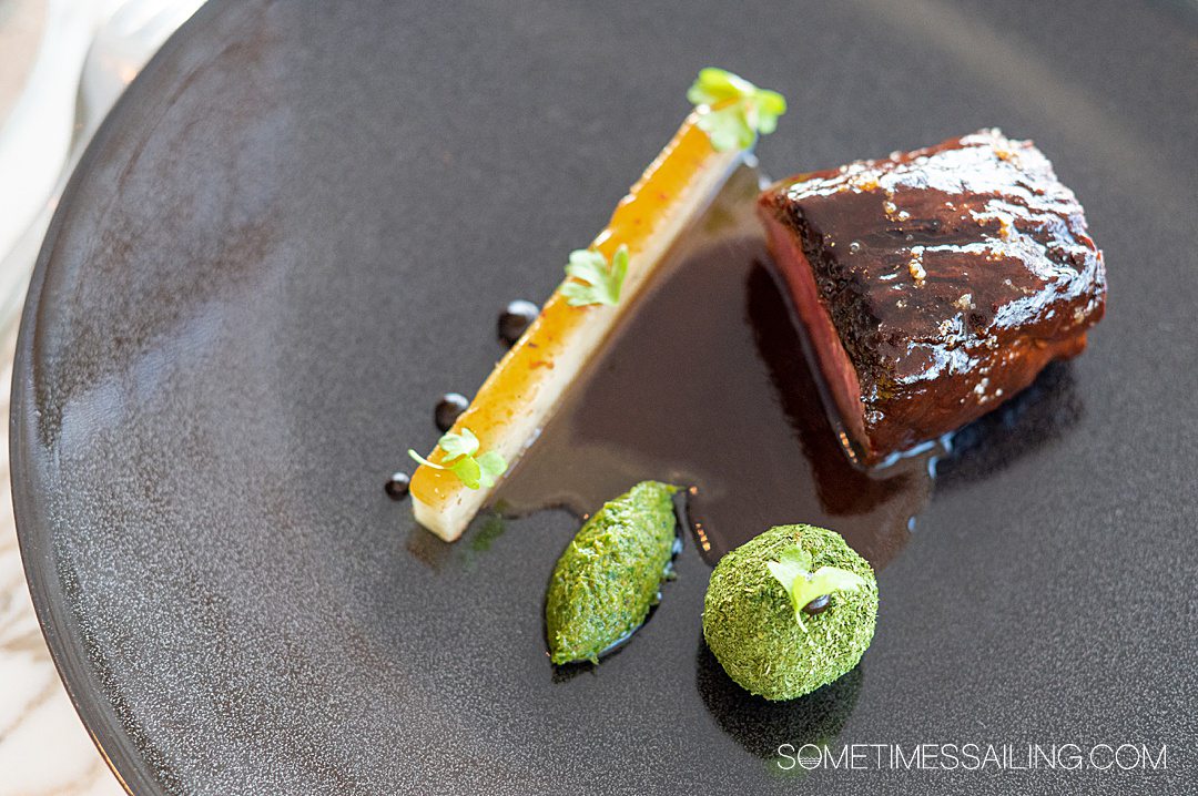 Black plate of food with a piece of beef on the right, a rectangular slice of a potato dish, and two green elements on the bottom at Enchante on Disney Wish cruise ship.