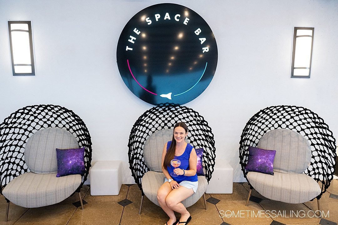 Woman sitting in a black net chair holding a cocktail under a round "The Space Bar" sign.