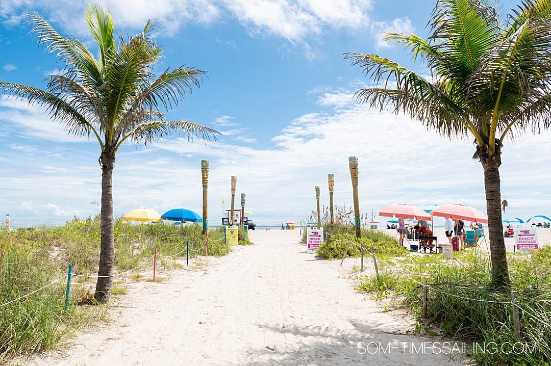 The sandy walkway leading to Cocoa Beach, with two palm trees on either side and a blue sky with white clouds.