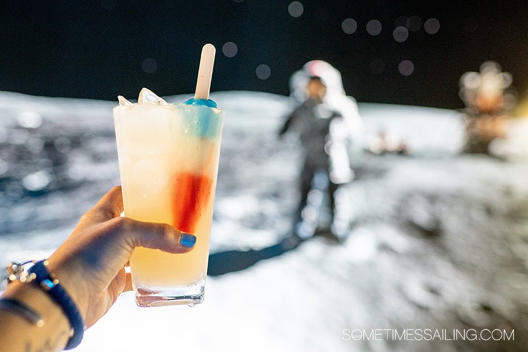 Hand holding a yellow drink in a pint glass with a Rocket Pop in it with a blurry photo of a man on the moon behind it at The Space Bar in Titusville near Port Canaveral.