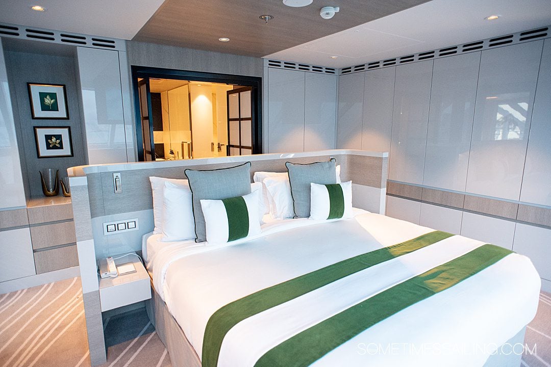 Bed with white and green striped bedding and door to the bathroom behind it in the Iconic Suite on Celebrity Beyond cruise ship.