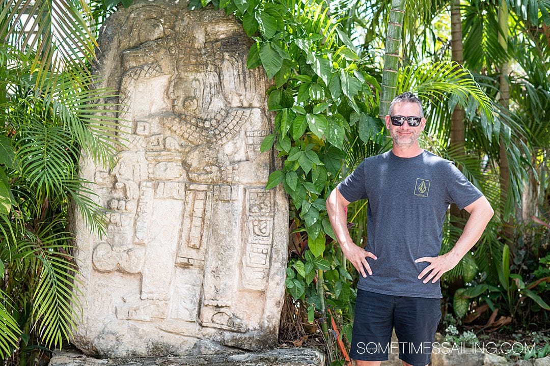 Man next to a Mayan ruin and a wall of lush palm plants behind him.
