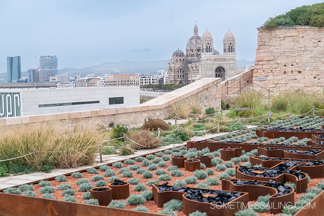 Aerial view of the city of Marseille as seen from the La Joliette Neighborhood, looking over a small garden, down at a large church, and to the cruise ships in the far distance.