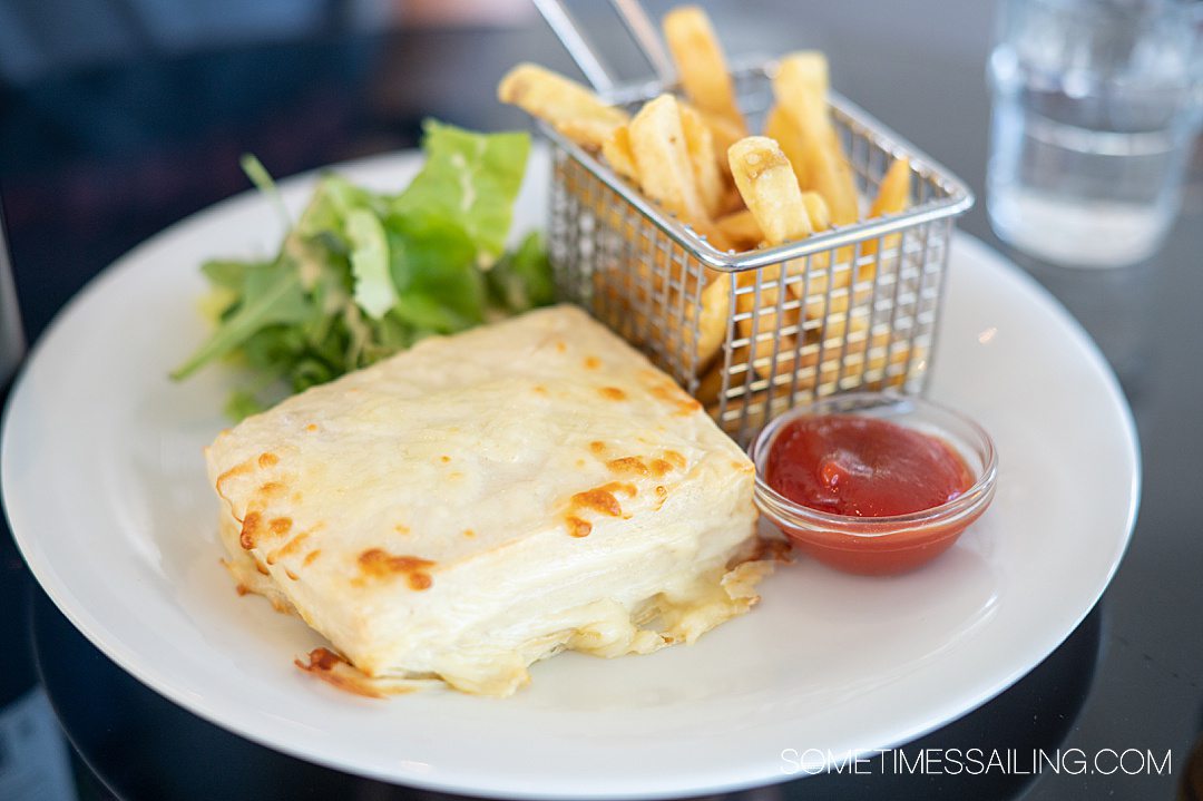 Melted cheese on a square Croque Monsieur sandwich with a mini basket of French fries, along with ketchup and salad.