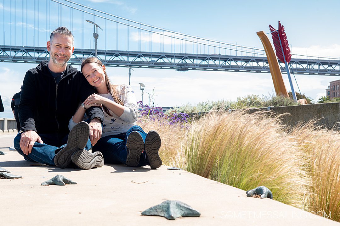 Couple in California with a bridge behind them and grass plants to the right.