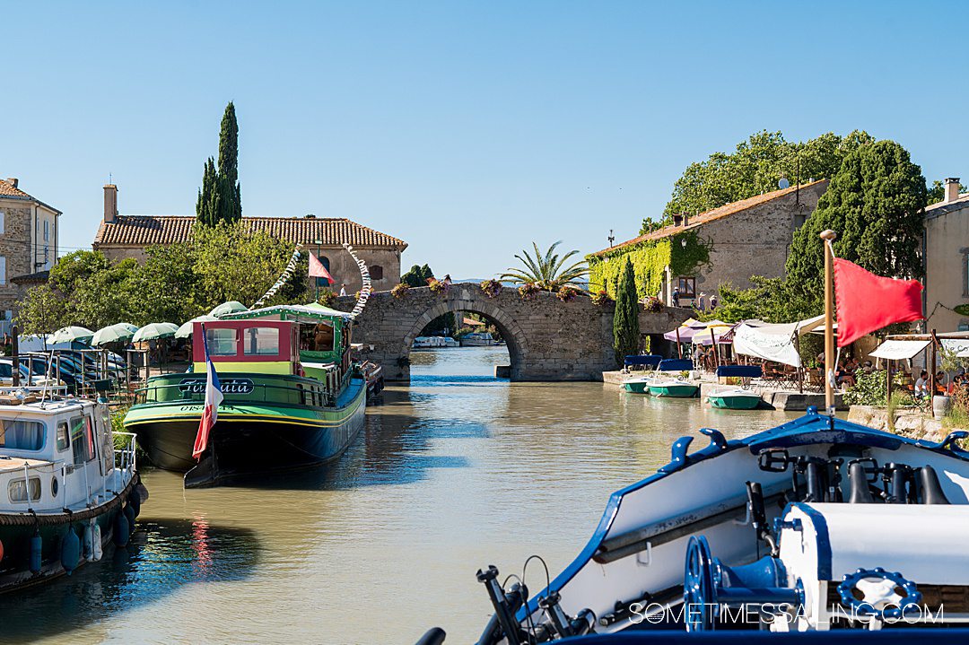 The bow of a boat sailing towards a bridge in France on a historic Canal with other boats lining the water.