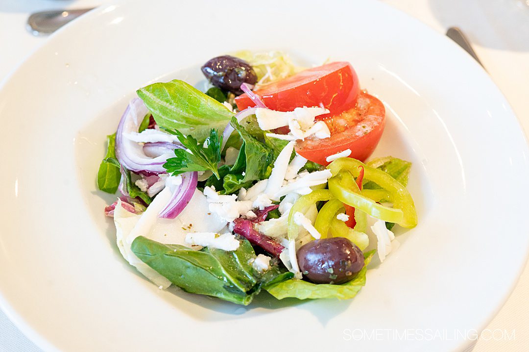 Salad in a white bowl with lettuce, tomato, red onion, Kalamata olives and cheese.