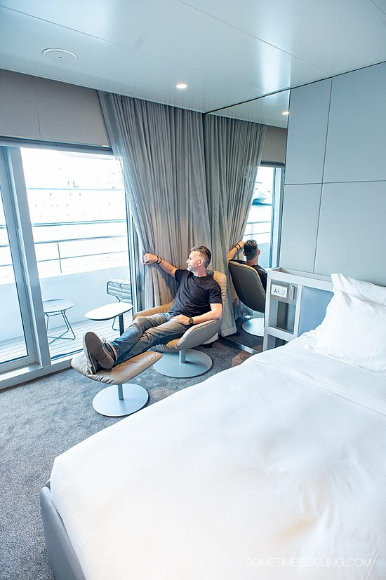 Inside a stateroom on Emerald Azzurra with a bed in white linens and man looking outside the windows.