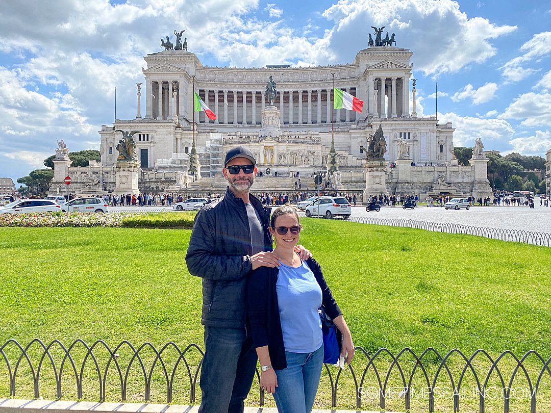 Couple in front of a monument in Piazza Venezia in Rome, Italy with Italian flags waving next to the landmark in the distance.