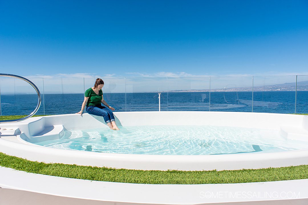 Woman sitting on the side of a hot tub on a luxury yacht with her feet in the water and blue ocean behind her.