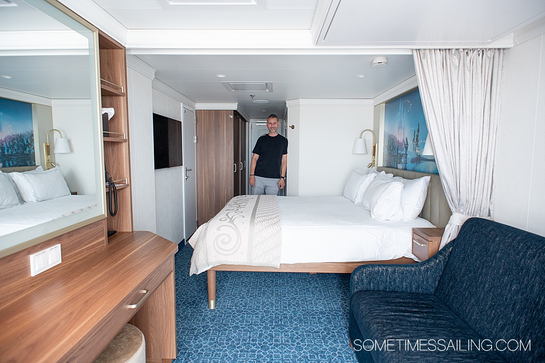 Inside a cabin stateroom on Disney Wish cruise ship, adults, only, with a man standing by the wall past the bed in the distance.