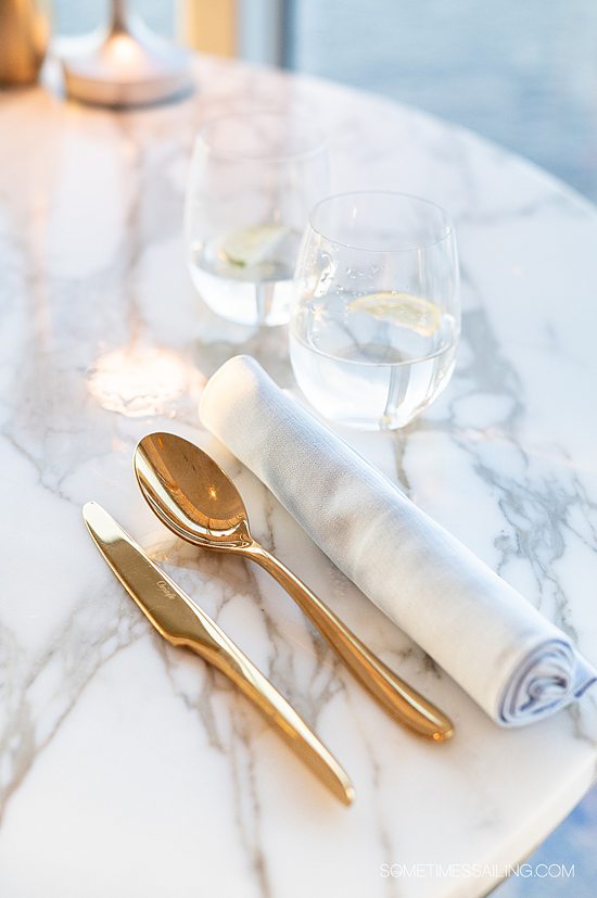 Gold spoon and butter knife next to a rolled napkin and two water glasses on a white marble table.