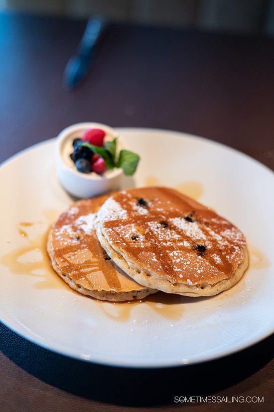 Two blueberry pancakes on a plate with powdered sugar and syrup drizzle and a small dish of fruit next to it.