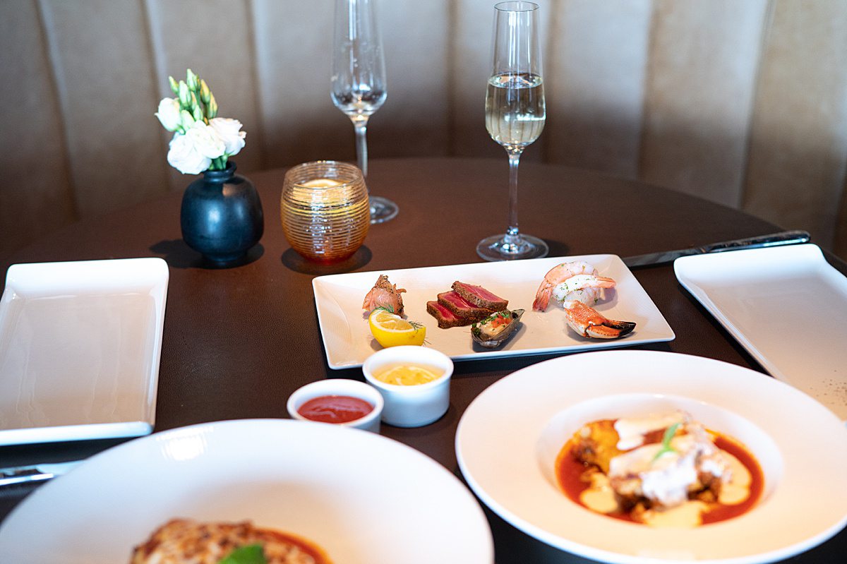 Table filled with food at Palo Italian restaurant on Disney Wish, with two glasses of champagne and a small black vase with flowers and a votive candle.