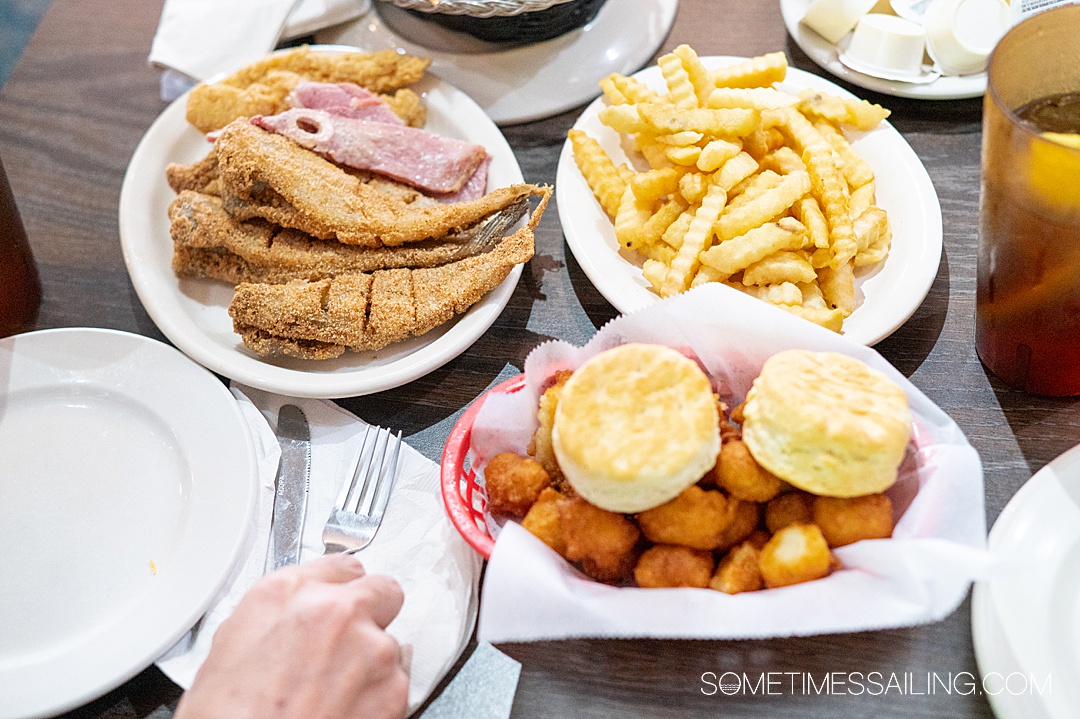 Table with plates of food: fried catfish, ham and chicken tenders, French fries, and a basket of hush puppies with two biscuits.