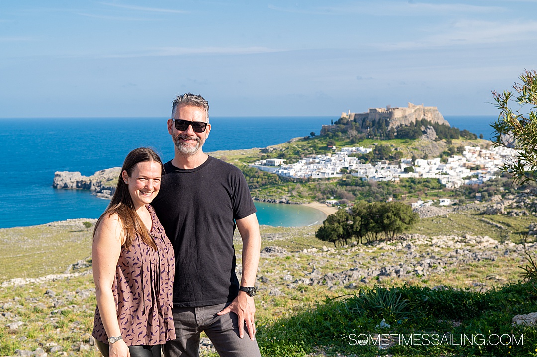 Woman and a man in front of a landscape view of Lindos, on the Greek island of Rhodes during a sunny day.