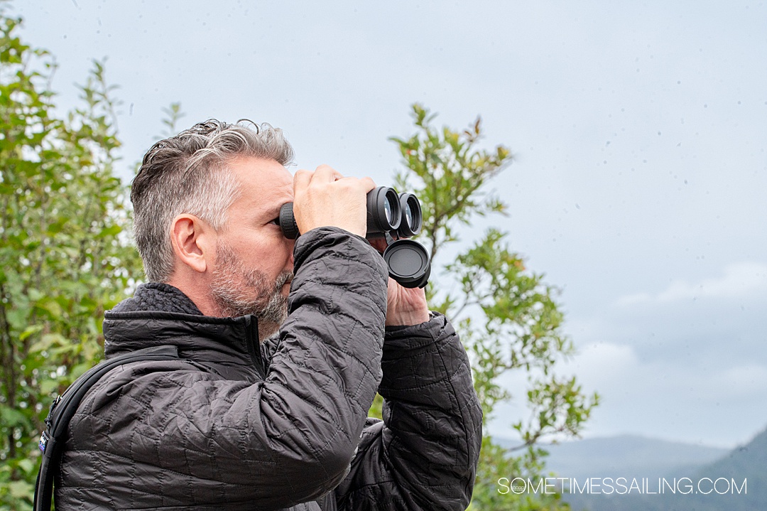 Man looking through binoculars with a black jacket and black hat on.