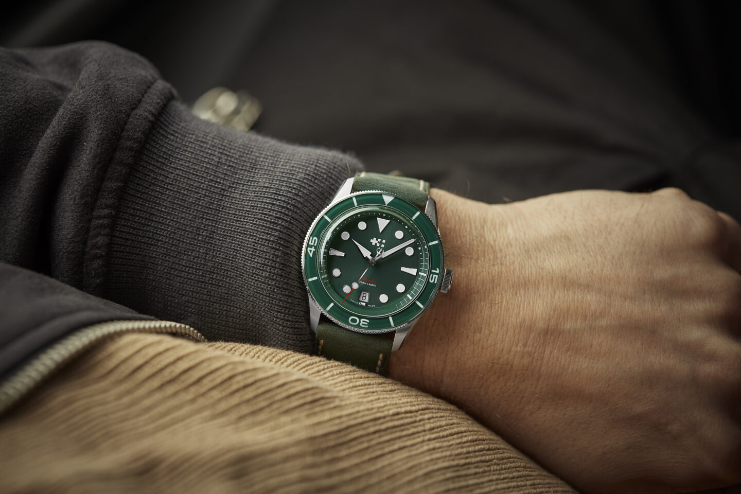 Green Christopher Ward watch on the wrist of a man wearing a long sleeve sweater. 