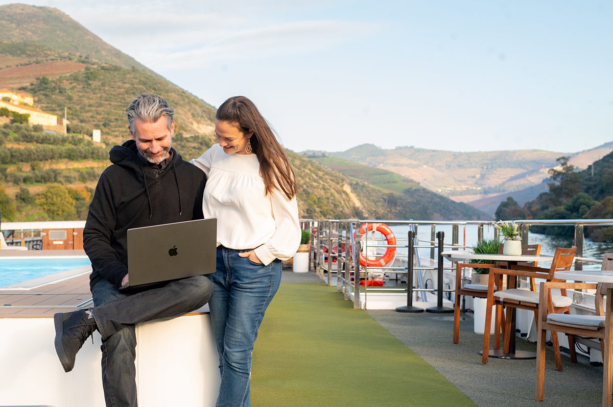 Man and a woman on the top deck of a river cruise ship sitting on the edge of pool with a computer in the man's lap and the mountains behind them.