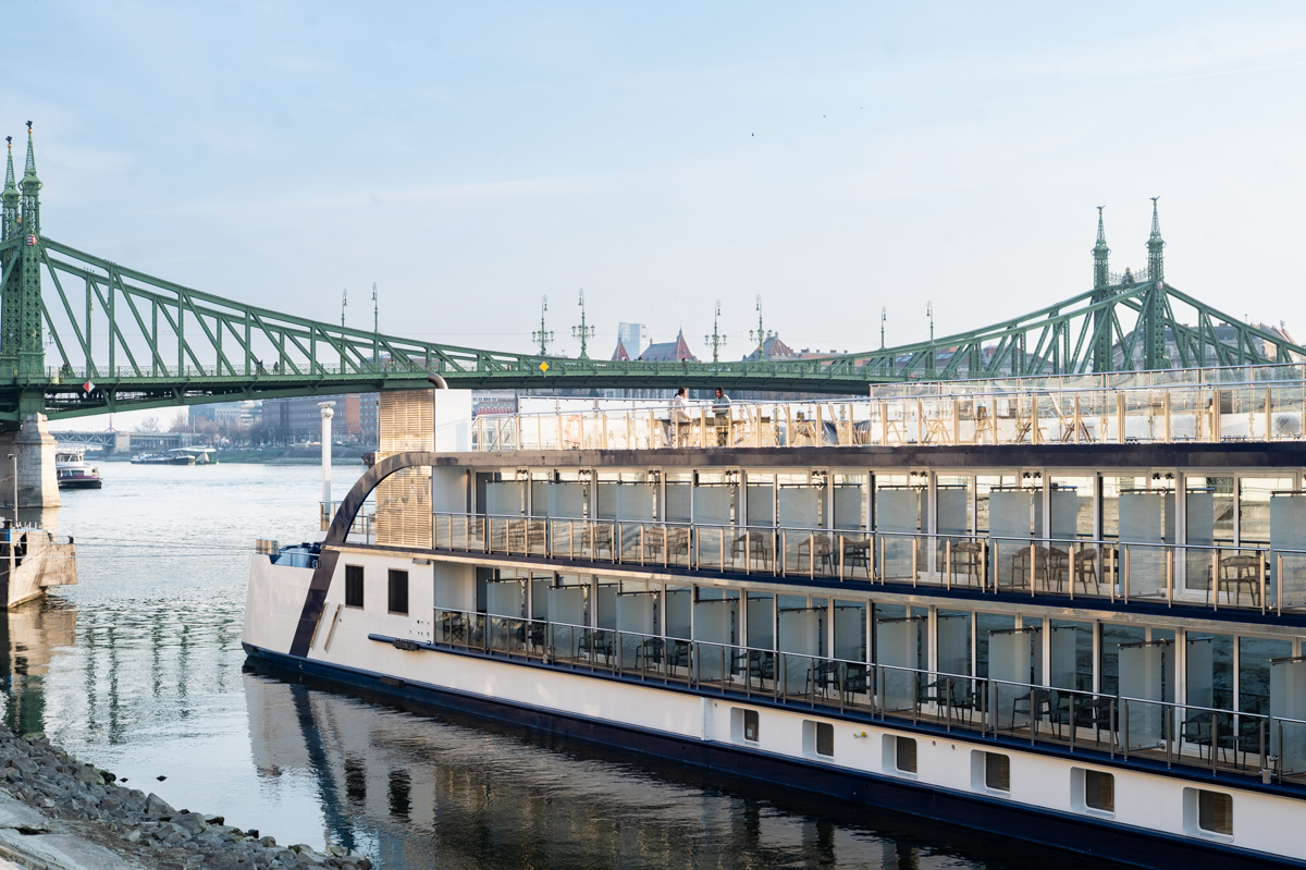 Exterior Starboard side of the AmaMagna river cruise ship with two decks of balconies and Budapest's Green Bridge in the background with a blue sky.