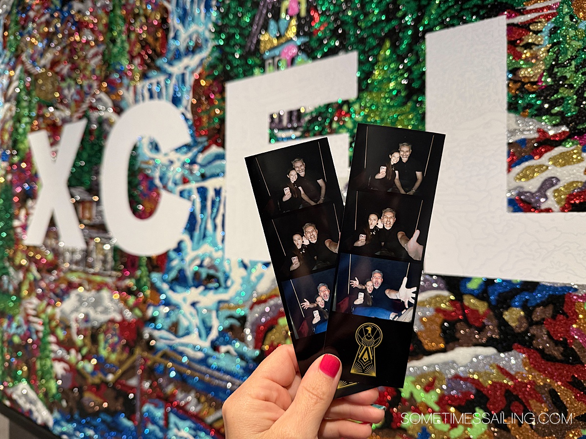Hand holding two photo strips in front of a glitter Xcel painting on the cruise ship.