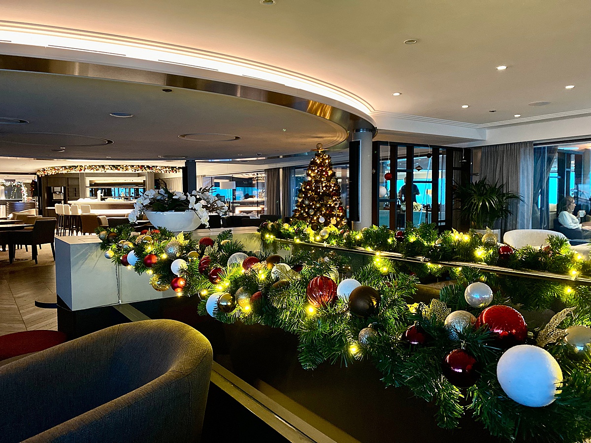 Holiday gardland lit up with white lights with red, white and gold ornaments and a Christmas tree in the distance on a river cruise ship.